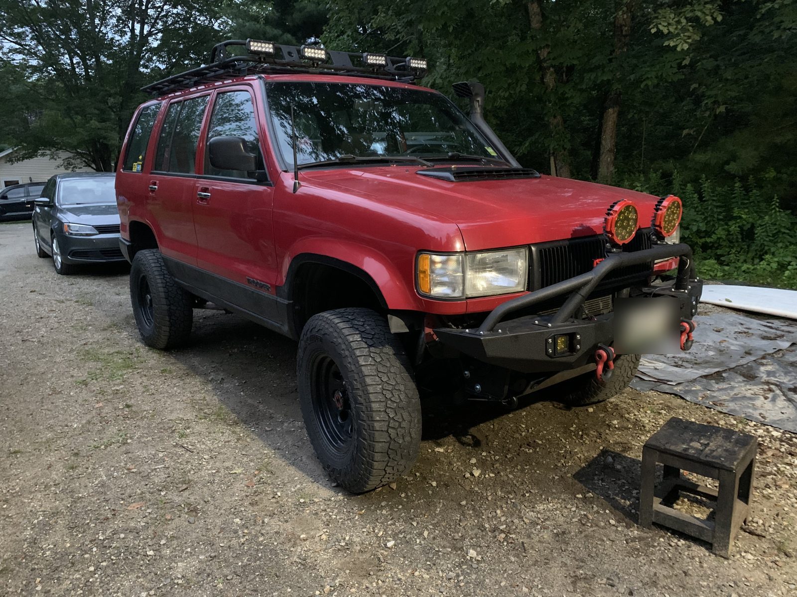 The red Isuzu Trooper a year later, bumper with red shackles and amber driving lights, parked on gravel in summer